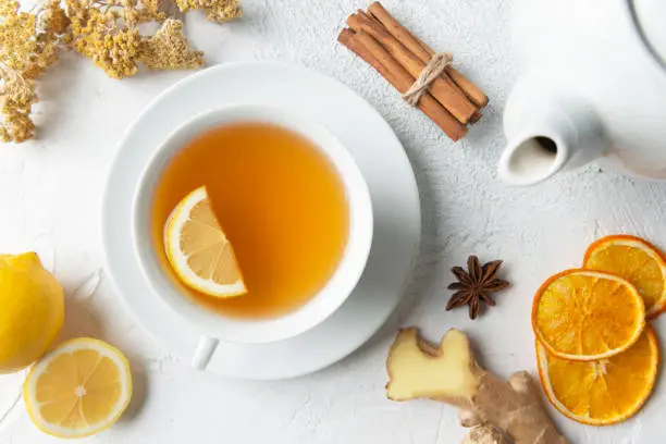 Directly above view of a tea cup with herbal tea and a slice of lemon in it. Surrounded by herbal tea ingredients like chamomile, chamomile tea, lemon, mint leaf, anise, ginger slice, dry orange slices and cinnamon.