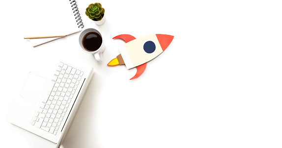 Directly above view of a laptop, notepad, model spaceship, flower and cup of coffee on pure white background. Representing desktop of a startup company.