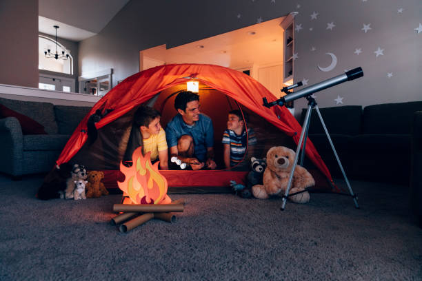 Father and Sons Camping Indoors A dad with his two sons camp inside their home due to the coronavirus restrictions and quarantine. They have pitched a tent along with stuffed animal friends and have a fake campfire next to a telescope. They are making the best of their situation and long to return to the outdoors. staycation stock pictures, royalty-free photos & images