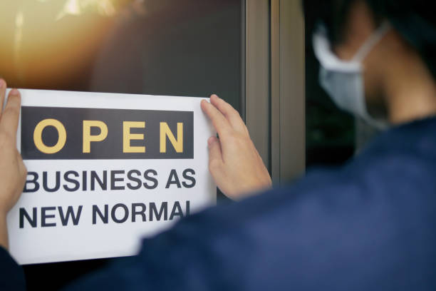 Reopening for business adapt to new normal in the novel Coronavirus COVID-19 pandemic. Rear view of business owner wearing medical mask placing open sign "OPEN BUSINESS AS NEW NORMAL" on front door. Reopening for business adapt to new normal in the novel Coronavirus COVID-19 pandemic. Rear view of business owner wearing medical mask placing open sign "OPEN BUSINESS AS NEW NORMAL" on front door. new normal concept stock pictures, royalty-free photos & images