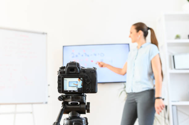 istock Online studying, online learning. Female teacher is recording classes on a camera, she stands near flip chart and screen with a marker in hand, camera in foreground 1223873375