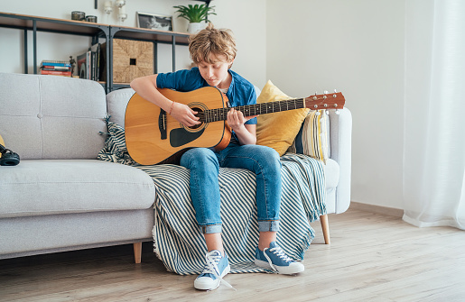 Preteen boy playing acoustic guitar dressed casual jeans, shirt and new sneakers sitting on the cozy sofa at home living room. Music education concept image.
