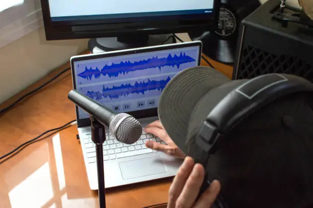 Photo of Person with cap and headphones recording a podcast at home using microphone and computers. Recording online podcast.