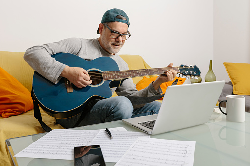 Musician playing the guitar and composing music with his colleagues by video conference through his laptop