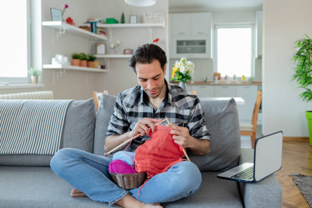 Knitting Tutorial Charming Man Watching Knitting Tutorial With Needles While Sitting on the sofa in the Living Room. Self-taught Caucasian Man Want to Learn New Hobby With Online Lessons in Laptop. Interesting Hobby for Mental Health knitting for beginners stock pictures, royalty-free photos & images