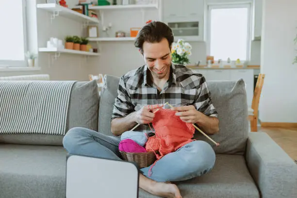 Charming Man Watching Knitting Tutorial With Needles While Sitting on the sofa in the Living Room. Self-taught Caucasian Man Want to Learn New Hobby With Online Lessons in Laptop. Interesting Hobby for Mental Health