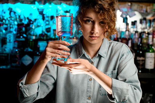 Pretty and young lady barman holds glass with fresh ready-made rose cocktail in her hands.