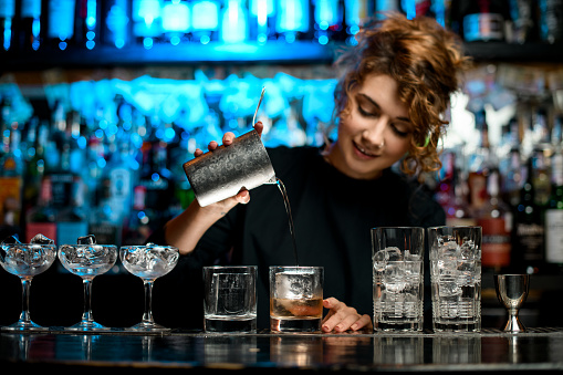 Lady barman carefully pours finished alcoholic cocktail into glass with ice. Different glasses stand on bar counter.