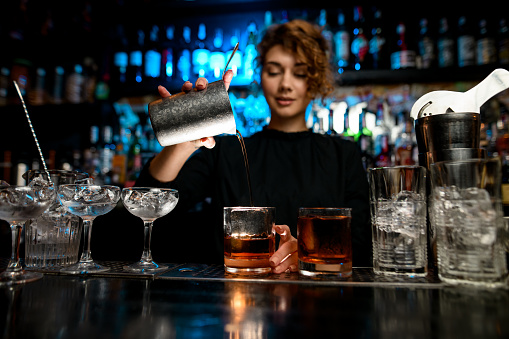 Young lady at bar pours alcoholic cocktail into glass. Different bar equipment stand on bar counter.