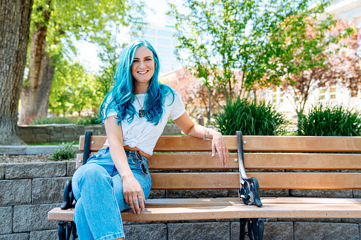 Unique Spunky Fashionable Young Woman with Fun Cute Teal Blue Green Dyed Hair Sitting on a Park Bench Outdoors in the Summer