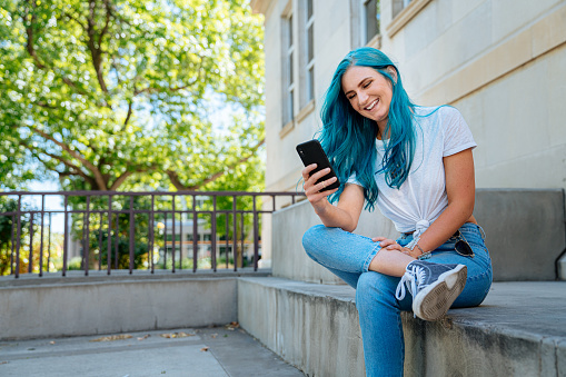 Unique Spunky Fashionable Young Woman with Fun Cute Teal Blue Green Dyed Hair Using Her Mobile Cell Phone to Text Friends, Check Her Email, Check Her Bank Account Balance, and Stay Connected Outdoors in the Summer