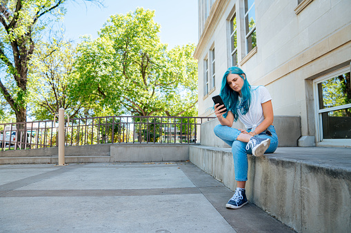 Unique Spunky Fashionable Young Woman with Fun Cute Teal Blue Green Dyed Hair Using Her Mobile Cell Phone to Text Friends, Check Her Email, Check Her Bank Account Balance, and Stay Connected Outdoors in the Summer