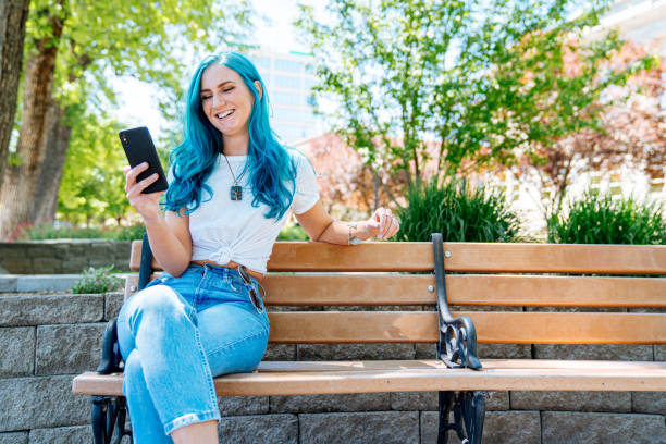 mulher jovem spunky elegante exclusiva com divertido teal azul azul cabelo coroudo verde usando seu smartphone celular para enviar mensagens de texto amigos, verificar seu e-mail, verificar seu saldo de conta bancária, e ficar conectado enquanto estiver a - blue hair - fotografias e filmes do acervo