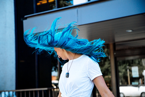 Artsy Low Angle Portrait Shot of a Beautiful, Unique Spunky Fashionable Young Woman with Fun Cute Teal Blue Green Dyed Hair Standing Flipping Her Hair in the Wind Outdoors in the Summer