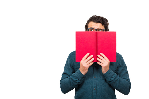 Student guy hiding behind a red book, looking suspicious aside isolated on white background with copy space. Scared nerd person, wearing eyeglasses, try to be invisible or incognito, covers face.