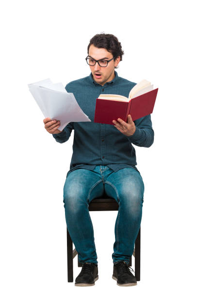 istock Full length of perplexed student looking stunned at paper projects and book in his hands, seated on chair isolated on white. Bewildered man wears glasses, looking confused at a textbook and sheets. 1223935413