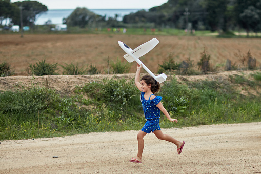 Side view of young girl wearing summertime casual clothing and smiling as she runs on dirt road holding toy glider overhead near Costa Brava beach.