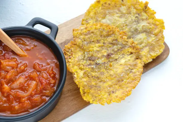 Photo of Fried banana with tomato sauce in a dark pot on a kitchen table with a white background. Latin cuisine concept. Patacón con hogao.