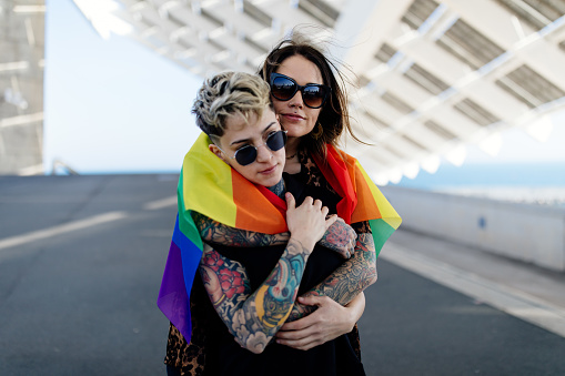 Alternative hipster lesbian couple embracing each other with LGBT pride rainbow flag, wearing glasses