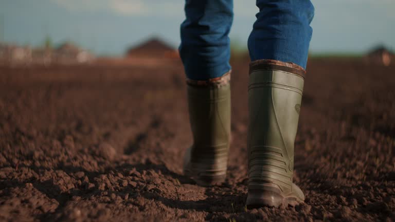 A farmer walks across a field in rubber boots on a blurred background of the tractor in motion. Concept of: Rubber boots, Lifestyle, Farmer, Slow Motion, Fields