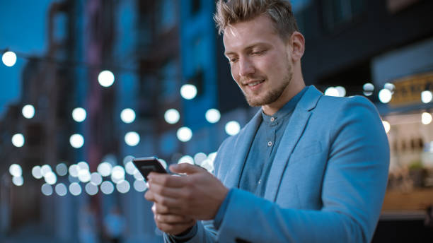 istock Handsome Young Concentrated Man is Using a Smartphone while Standing Outdoors in the Evening. He's Browsing the Internet or Social Media, Posting a Status Update. Man is Wearing a Suit. 1223977317