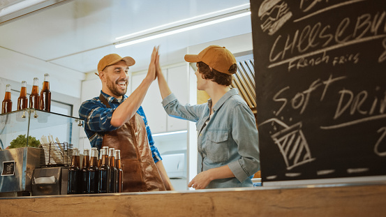 Successful Food Truck Male and Female Employees Give High Five and Celebrate. They Wear Brown Caps. They are Cheerful and Smiling. Commercial Truck or Kiosk Selling Street Food and Drinks.