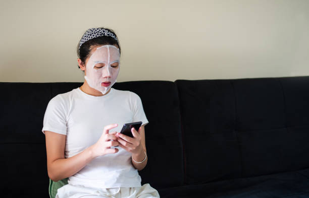 istock Asian woman using phone while applying cosmetic face mask at home 1223980347