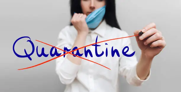 Quarantine End Concept. A female hand in a white smock holds a red pen and crosses out the handwritten word Quarantine. Gray background. Take off mask. Coronavirus, COVID-19.