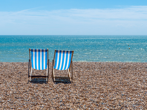 Two blue and white deck chairs on a shingle beach on a sunny day with blue sky.