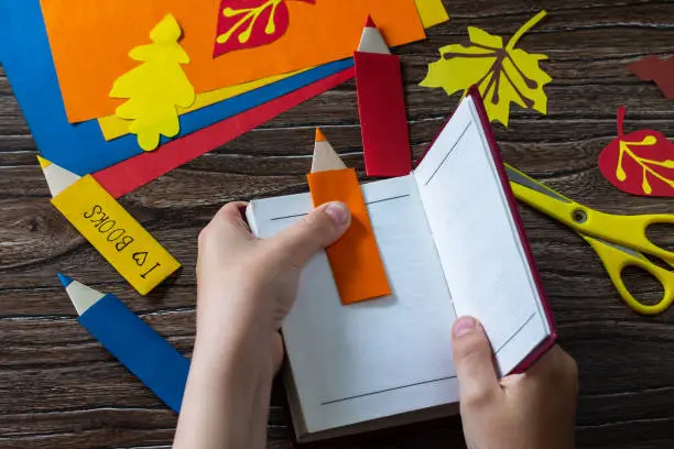 Photo of Child holds multi-colored bookmarks origami pencil made of paper new school year on a wooden table. Welcome to school. Children's art project, needlework, crafts for children.