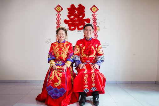 An Asia couple having their Chinese traditional wedding ceremony. They sitting together in traditional clothes. Just married. Hands on knees. Smiling