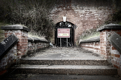 A danger sign at the entrance to a tunnel, reached by a flight of steps. The sign reads “Danger! Do Not Enter. Warning. Deep Excavations.” Subdued colour.