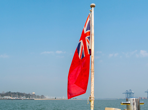 A Red Ensign being flown from the stern of a small boat in the Orwell-Stour estuary between, Shotley and Felixstowe in Suffolk, and Harwich in Essex. Some of the cranes of Port of Felixstowe can be seen on the right while the masts of yachts at Shotley Gate Marina can be seen on the left.