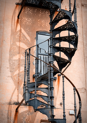 An ornate wrought iron spiral staircase on the outside of a tall building, with shadow.