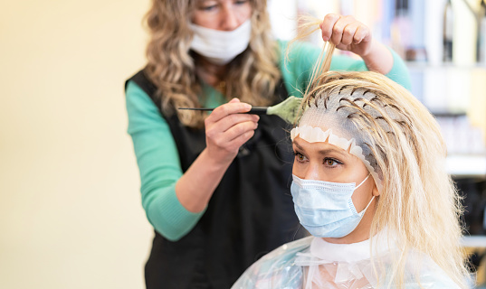 Hairdresser bleaching strands of blonde woman hair with a rubber hat in beauty salon, during COVID-19