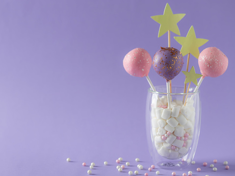 pastel colored cake pops in a glass with marshmallows and sprinkles. birthday festive dessert. purple background. horizontal image. place for text.