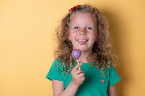 Happy adorable girl shows tongue and eats tasty cake pops, candy on a stick. happy childhood. yellow background