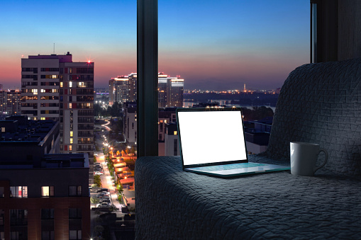 Opened laptop on grey sofa with panoramic view window of night city