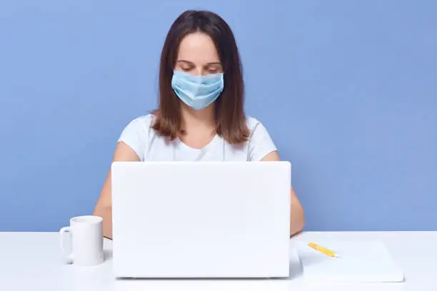 Horizontal shot of dark haired lady wearing casual t shirt and medical protective mask working in front of lap top screen, looks concentrated, student female doing online university task, education.