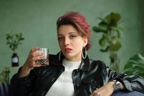 Photo of Young sad woman with dark pink hair with glam rock style look is drinking whisky from a glass on green background in studio, woman's alcoholism, alcohol addict concept