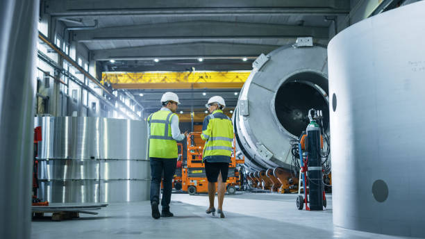 istock Two Heavy Industry Engineers Walk Through Pipe Manufacturing Factory, Use Digital Tablet Computer, Have Discussion. Facility for Construction of Oil, Gas and Fuel Pipeline Transportation Products 1224034099