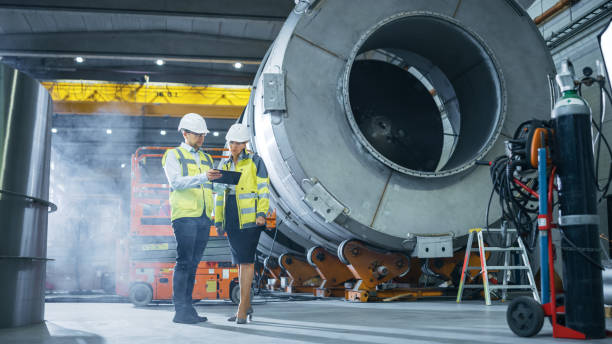 istock Two Heavy Industry Engineers Stand in Pipe Manufacturing Factory, Use Digital Tablet Computer, Have Discussion. Facility for Construction of Oil, Gas and Fuel Pipeline Transportation Products 1224034108
