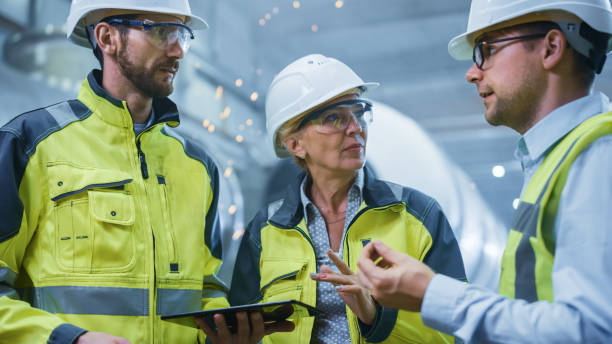 istock Three Heavy Industry Engineers Stand in Pipe Manufacturing Factory, Use Digital Tablet Computer, Have Discussion. Large Pipe Assembled. Design and Construction of Oil, Gas and Fuels Transport Pipeline 1224034111