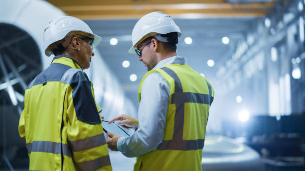 istock Two Heavy Industry Engineers Stand in Pipe Manufacturing Factory, Use Digital Tablet Computer, Have Discussion. Construction of Oil, Gas and Fuels Transport Pipeline. Back View Sparks Flying 1224034136