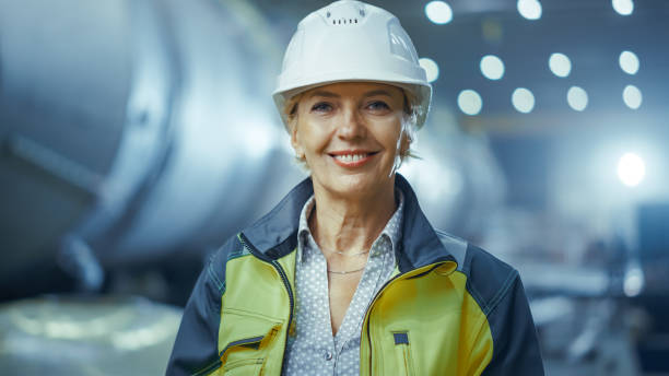 portrait de l’ingénieur féminin professionnel d’industrie lourde portant l’uniforme de sécurité et le chapeau dur, souriant charmant. dans le fond une grande usine industrielle non ciblée où les étincelles de soudage volant - charmingly photos et images de collection