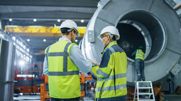 istock Two Heavy Industry Engineers Walk Through Pipe Manufacturing Factory, Use Digital Tablet Computer, Have Discussion. Facility for Construction of Oil, Gas and Fuel Pipeline Transportation Products 1224034767