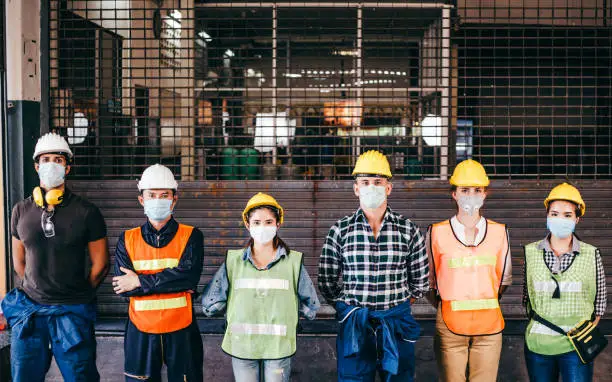 Photo of Group of industrial or engineer corporate workers wear protective mask and hard hat helmet standing line up in front of factory lock down prevention for Coronavirus or COVID-19 epidemic outbreak