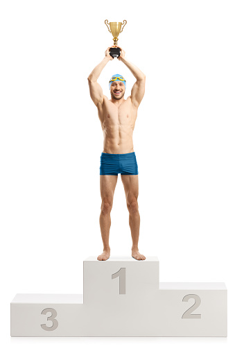 Full length portrait of a male swimmer lifting a gold trophy cup on a winners podium isolated on white background