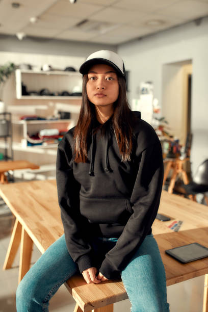 Creative streetwear. Cheerful female worker looking at camera while posing in custom apparel, baseball cap and hoodie. Young woman working at custom T-shirt, clothing printing company