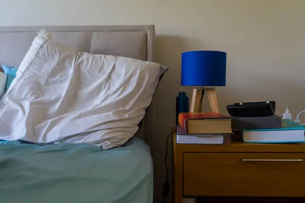 Photo of an unmade bed and bedside table with books and a blue lamp